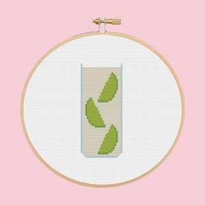 May include: A white embroidery hoop with a cross-stitch design of a glass of lime juice. The glass is filled with a light green liquid and has three lime wedges inside.