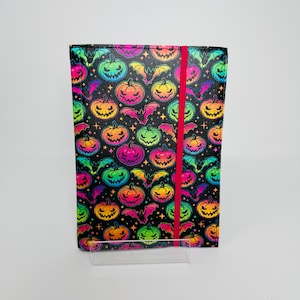 May include: A Halloween-themed journal with a black cover featuring a vibrant pattern of colourful jack-o'-lanterns and bats. A bright red elastic band secures the journal. The journal is displayed on a clear acrylic stand.
