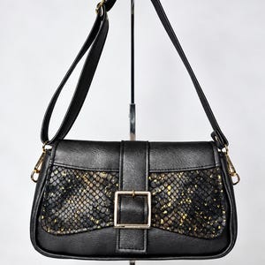 Può includere: Una borsa a tracolla nera con un pannello testurizzato a motivo pelle di serpente con macchie dorate. La borsa include un cinturino verticale centrale con una fibbia rettangolare dorata, una tracolla regolabile abbinata e finiture in metallo dorato.