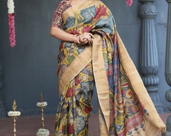 May include: A woman wearing a beige and blue silk saree with a floral print. The saree has a gold border and is draped over a blouse with a black and red print. The woman is standing in front of a gray wall with a garland of white flowers hanging on it.