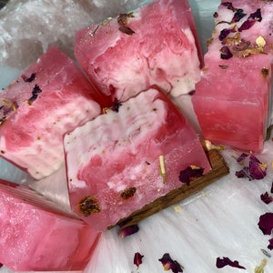 May include: Pink and white swirled soap bars with dried rose petals and cinnamon sticks. The soap has a translucent quality, with a marbled effect. The bars are arranged on a white, fluffy surface, with scattered rose petals.