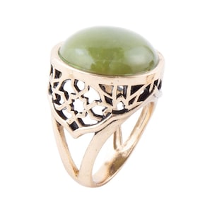 May include: A gold-toned ring featuring a large, oval, olive-green stone. The ring's band has an openwork design with geometric shapes. The ring is set against a white background, highlighting its details and color.
