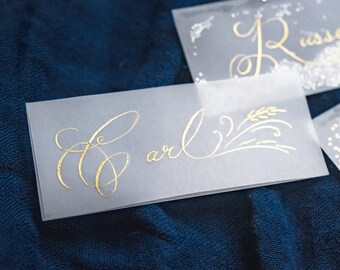 Può includere: Tre segnaposto traslucidi con calligrafia dorata. Le carte sono su uno sfondo di tessuto blu scuro. I nomi sulle carte sono "Carl", "Russell" e un nome illeggibile.