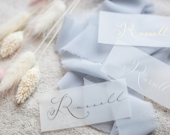 Può includere: Tre segnaposto di carta bianca con il nome "Russell" scritto in calligrafia. Le carte sono su un tessuto blu chiaro.