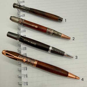 May include: Four ballpoint pens arranged diagonally on lined paper, labeled 1 through 4. Each pen features a wooden barrel combined with metal components in gunmetal, copper, or silver tones, showcasing various intricate engraved patterns.