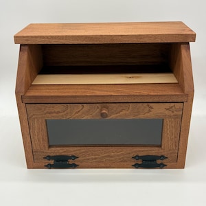 May include: A wooden bread box with a dark stain finish. The box has a hinged door with a glass window and decorative black metal hinges. The top of the box has a small shelf. The interior has a light wood shelf.