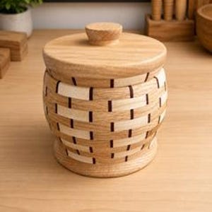 Può includere: Un contenitore rotondo in legno con coperchio, caratterizzato da un motivo esterno intrecciato creato con strisce di legno alternate in tonalità chiare e medie e accenti verticali scuri. Il contenitore ha una base piatta e un coperchio circolare.