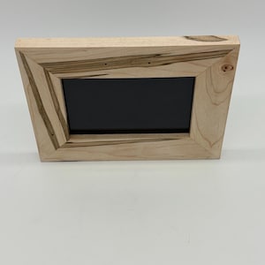 May include: A wooden picture frame with a black interior. The frame is made of light-colored wood with darker grain patterns and a unique angled design. The frame is rectangular and stands upright.