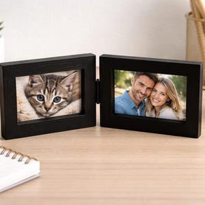 May include: A black double-hinged picture frame featuring two rectangular openings. The left frame displays a photograph of a tabby cat, while the right frame holds a photograph of a smiling couple. The frame is constructed with a smooth finish.