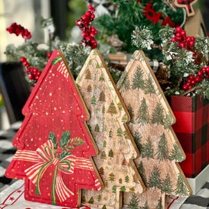 Puede incluir: Tres adornos de árbol de Navidad de madera. El primero es rojo con un diseño de regalo, el segundo tiene un patrón de árbol de Navidad y el tercero tiene un diseño de árbol verde. Los adornos están sobre una superficie a cuadros blancos y negros.