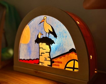 May include: A wooden nightlight shaped like a half-circle with a stained glass design of a house, a chimney, and two storks. The nightlight is lit from behind, casting a warm glow.