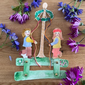May include: Two children, one wearing a blue shirt and the other wearing a yellow shirt, are holding strings attached to a maypole decorated with a green wreath. The maypole is standing on a green bench with flowers painted on it. The background is a wooden surface with purple and blue flowers.