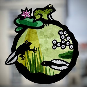 May include: A whimsical paper cutout depicting a pond scene with a frog, tadpoles, and lily pads. The frog is green with black spots, and the tadpoles are black and white. The lily pads are green with a pink flower.