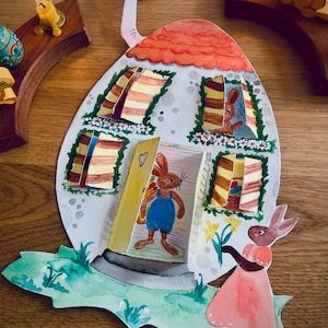 Könnte beinhalten: Ein Osterhasenhaus aus Papier mit rotem Dach, gelben Fenstern und einem braunen Hasen, der aus der Tür schaut. Ein zweiter Hase in einem rosa Kleid hält einen Strauß gelber Blumen vor dem Haus.