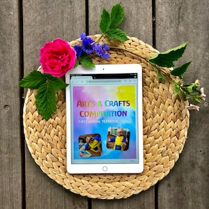 以下が含まれることがあります： 白のタブレットコンピュータに、芸術と工芸の年鑑のカラフルな表紙が表示されています。表紙には「Arts & Crafts Compilation」というタイトルと「First Annual Yearbook, 2020」というテキストが書かれています。