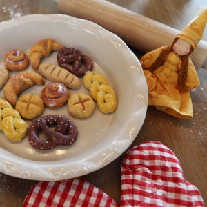Pode incluir: Um prato de torta branco cheio de vários produtos assados, incluindo pretzels, pães e pão trançado. O prato está sobre uma mesa de madeira com um descanso de panela vermelho e branco ao lado.