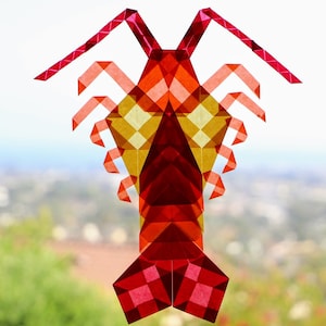 May include: A colorful, geometric papercraft of a shrimp. The shrimp is made of translucent red, orange, and yellow paper, with a detailed, angular design. The background is a blurred view of a green landscape.
