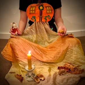 May include: A felt pumpkin house with a door and windows, a small white mouse figurine, a lit candle, and scattered autumn leaves on a yellow and orange fabric.