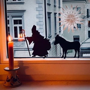 Puede incluir: Una silueta de una figura sosteniendo un bastón, un burro y una vela encendida en una ventana. La figura lleva una túnica y un sombrero. El burro está de pie junto a la figura. La vela está en un portavelas en un alféizar de la ventana.