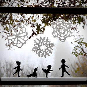 May include: Four paper snowflake cutouts are displayed on a window, with three black silhouette figures of people standing in front of them. The figures are facing forward with their arms at their sides.