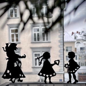 May include: Black silhouette cutouts of three figures, two girls and a woman, holding a heart and a star. The figures are set against a window background.