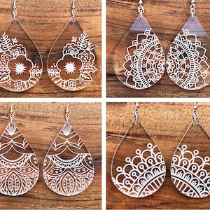 May include: Set of six white acrylic teardrop earrings with various floral and mandala designs. The earrings are on a wooden background.