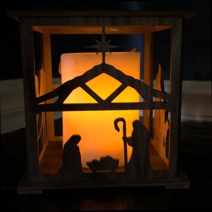 May include: A wooden candle lantern with a cutout design of the nativity scene. The candle is lit and glows through the cutout, creating a warm, inviting glow.