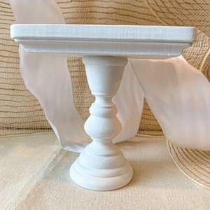 May include: White cake stand with a square top and a pedestal base. The stand is made of wood and has a smooth, painted finish.