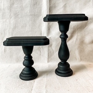May include: Two black wooden pedestal stands with a rectangular top. The stands are different sizes and are painted black.