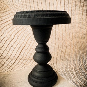 May include: Black wooden pedestal with a round top and a fluted base. The pedestal is on a beige textured surface.