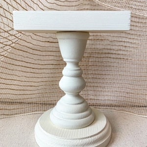 May include: White wooden cake stand with a round base and a square top. The stand is made of unfinished wood and has a simple, elegant design.