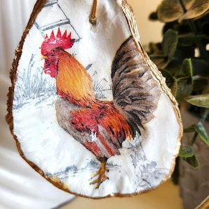 May include: A decorative oyster shell ornament featuring a colorful rooster illustration with red, orange, and black plumage. The shell has a metallic gold painted edge and is suspended by a thin gold cord, creating a hanging display piece.