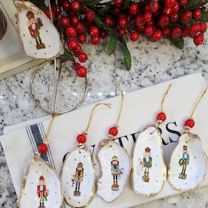 May include: Six handmade Christmas ornaments featuring nutcracker designs painted on oyster shells. Each ornament has a gold trim and a red bead on the string.
