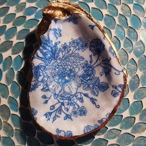 May include: A decorative oyster shell dish with a gold-painted rim and a white interior featuring a blue floral design. The shell is set on a mosaic tile surface with blue and teal tiles.
