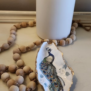 May include: A white oyster shell dish with a gold rim, featuring a hand-painted peacock design. The dish is on a white surface with a wooden bead garland.