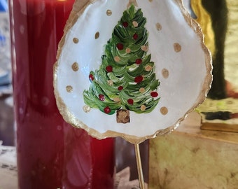 May include: A hand-painted oyster shell ornament featuring a green Christmas tree with red and gold ornaments on a white background. The shell has a gold-painted edge and hangs from a gold string on a thin metal display stand.