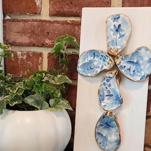 May include: A white wooden cross decorated with blue floral patterned oyster shells with gold trim. The cross is displayed against a brick wall, with a white pumpkin-shaped planter holding greenery in the foreground.