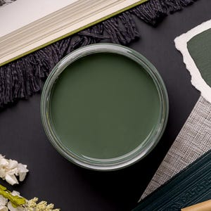 May include: An overhead shot of a jar filled with olive green paint. The jar is surrounded by a book with a green fringe, a small notebook, a pen, and white flowers on a black surface.