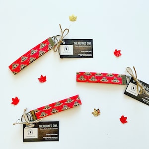 May include: Three red and brown keychains with a pattern of small brown hats. Each keychain has a silver key ring and a small brown tag with the text "The Refined Owl" and a small owl logo.