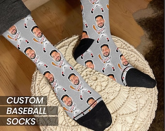 Puede incluir: Un par de calcetines de béisbol personalizados grises con un patrón repetido de la cara de una persona que lleva un uniforme de béisbol y sostiene un bate. El texto "CUSTOM BASEBALL SOCKS" está impreso en la parte inferior de la imagen.