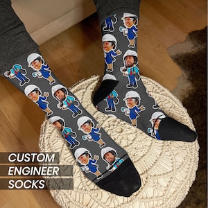 May include: A pair of grey socks with a custom design featuring multiple images of people wearing hard hats. The socks have black toes and heels. The text "CUSTOM ENGINEER SOCKS" is visible at the bottom of the image.