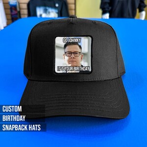 May include: Black snapback hat with a rectangular patch featuring a man wearing glasses and the text "GO JOHNNY IT'S YOUR BIRTHDAY." The hat also has the text "CUSTOM BIRTHDAY SNAPBACK HATS" on the brim.