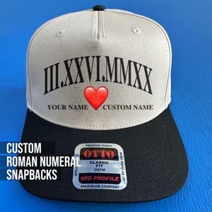 May include: A custom snapback hat with a black brim and a light gray crown. The hat features Roman numerals, a red heart, and space for custom names. The text "CUSTOM ROMAN NUMERAL SNAPBACKS" is on the side.