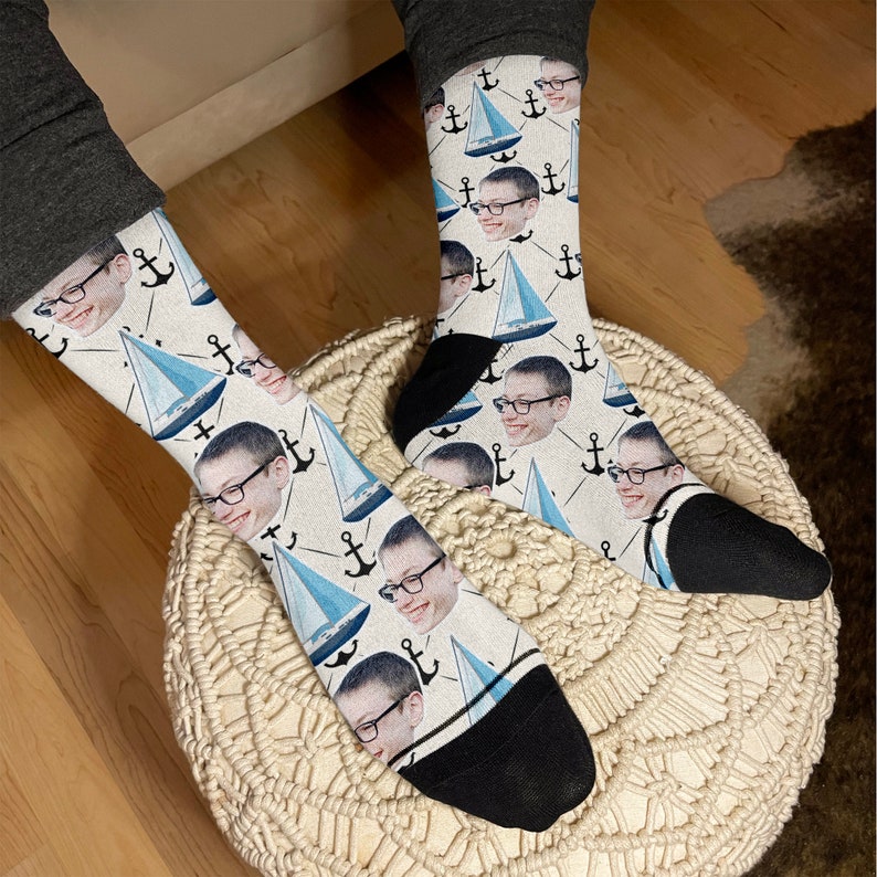 May include: A pair of white socks with a blue and white sailboat pattern. The socks feature a repeated image of a person's face with a black anchor.