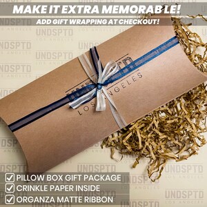 May include: A brown pillow box with a blue and white ribbon tied around it. The box is filled with brown crinkle paper. The box has the text "UNDSPTD LOS ANGELES" printed on it. The text "MAKE IT EXTRA MEMORABLE! ADD GIFT WRAPPING AT CHECKOUT!" is printed at the top of the image. A checklist with the text "PILLOW BOX GIFT PACKAGE", "CRINKLE PAPER INSIDE", and "ORGANZA MATTE RIBBON" is at the bottom of the image.