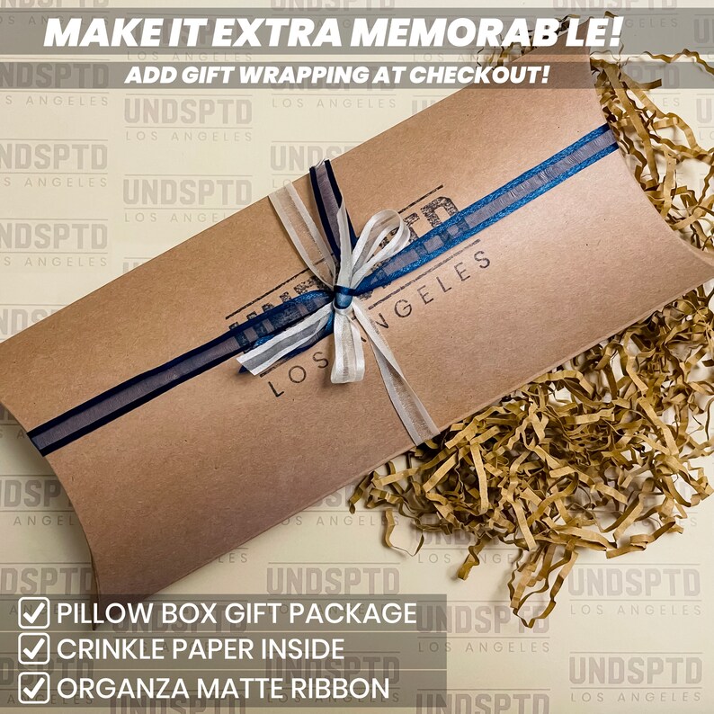 May include: A brown pillow box with a blue and white ribbon tied around it. The box is filled with brown crinkle paper. The box has the text "UNDSPTD LOS ANGELES" printed on it. The text "MAKE IT EXTRA MEMORABLE! ADD GIFT WRAPPING AT CHECKOUT!" is printed at the top of the image. A checklist with the text "PILLOW BOX GIFT PACKAGE", "CRINKLE PAPER INSIDE", and "ORGANZA MATTE RIBBON" is at the bottom of the image.