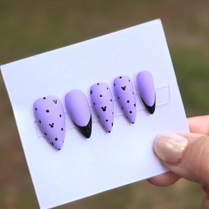 May include: Five almond-shaped press-on nails in a matte lavender finish. The nails feature black polka dots and small black mouse-ear silhouettes. Two nails have a solid black French tip design, while the others are covered in the dot pattern.