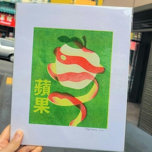 May include: Print of a stylized apple with a peeled spiral, set against a green background. The apple features red and white stripes, with the Chinese characters for "apple" in yellow. The print is held in a clear plastic sleeve.