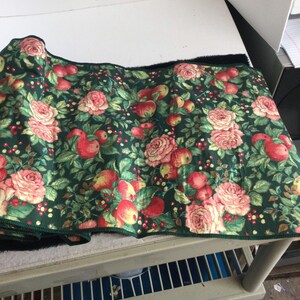 K&ouml;nnte beinhalten: Ein aufgerollter Stoff mit dunkelgr&uuml;nem Hintergrund, verziert mit einem floralen Muster aus roten &Auml;pfeln, rosa Rosen und kleinen roten Beeren. Der Stoff hat einen schwarzen Rand und einen gr&uuml;n gen&auml;hten Rand.