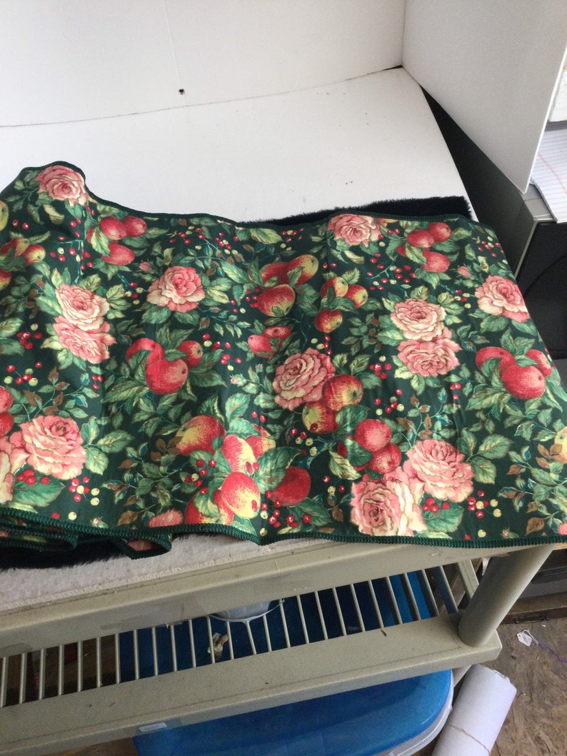 K&ouml;nnte beinhalten: Ein aufgerollter Stoff mit dunkelgr&uuml;nem Hintergrund, verziert mit einem floralen Muster aus roten &Auml;pfeln, rosa Rosen und kleinen roten Beeren. Der Stoff hat einen schwarzen Rand und einen gr&uuml;n gen&auml;hten Rand.